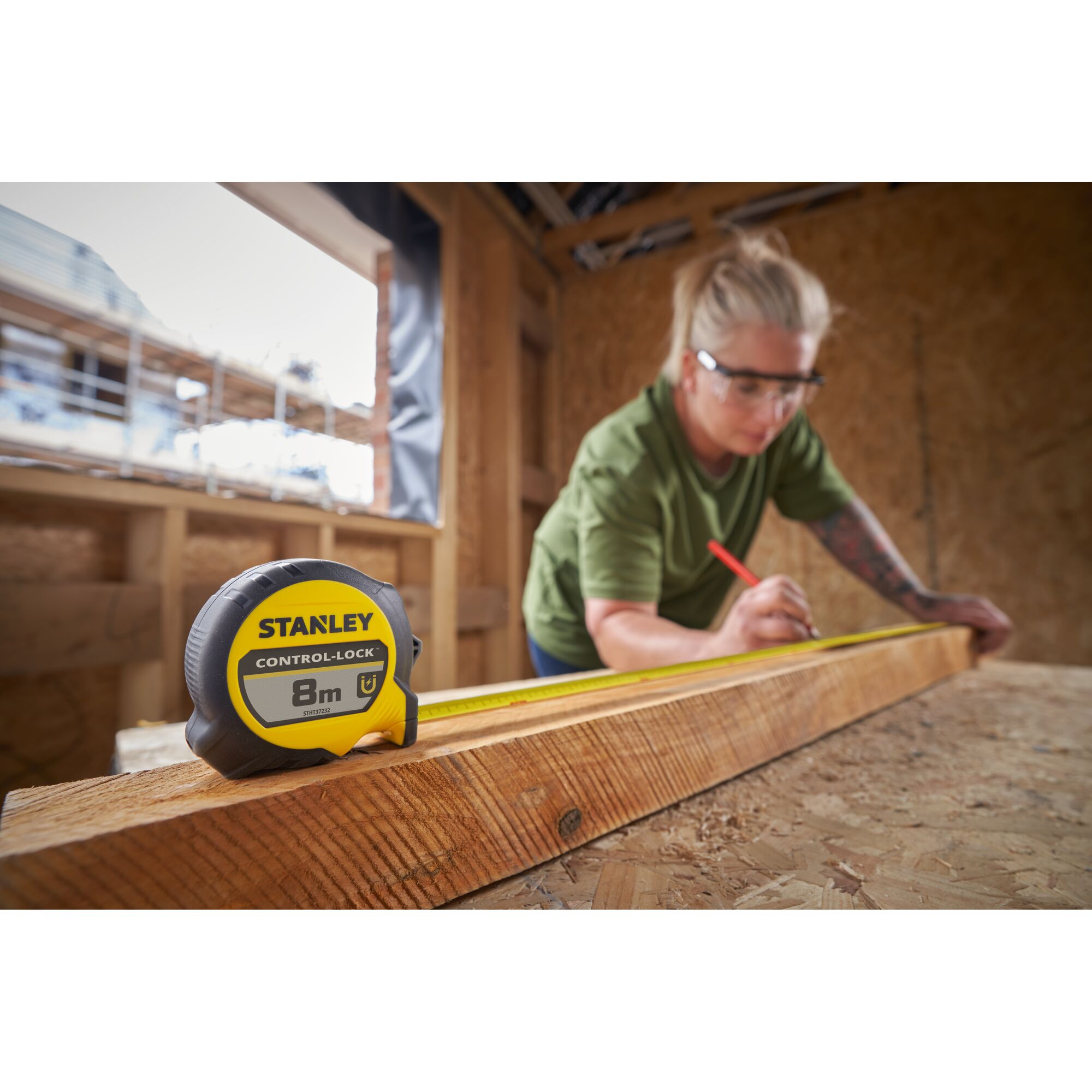 STANLEY STHT37232-0 tape measure on wooden beam, person marking measurements, indoor construction site.
